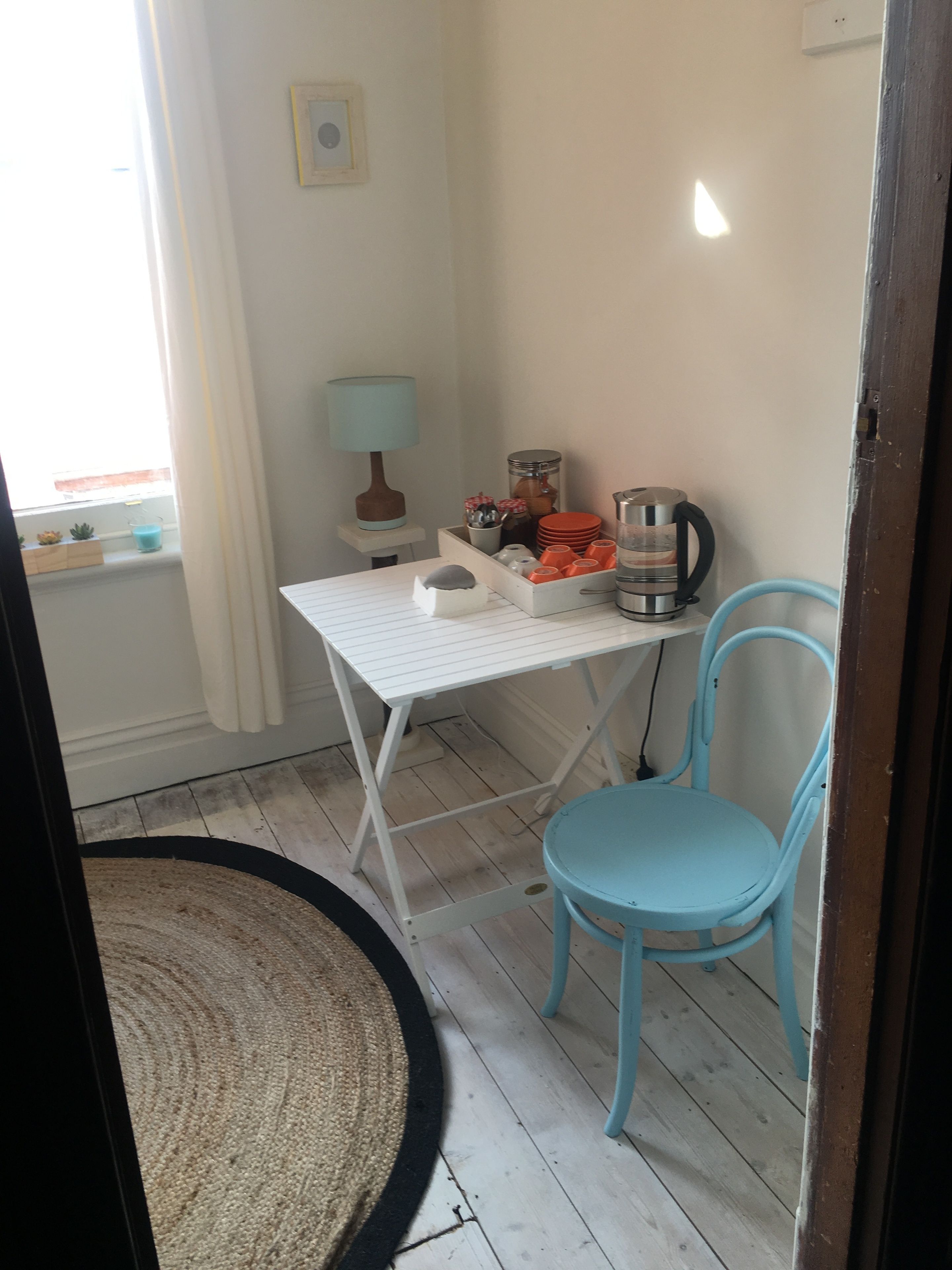 A small, well-lit room with a white table, a blue chair, a coffee maker, and a lamp.