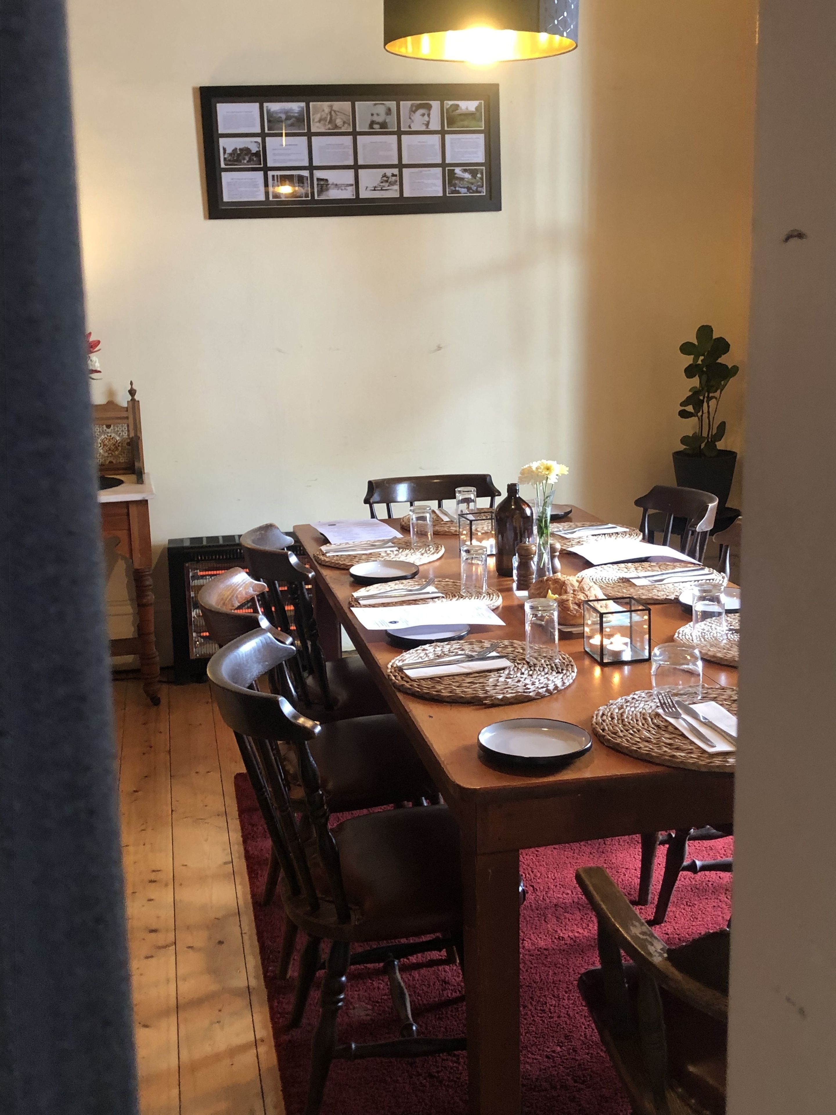 Comedor con una larga mesa de madera dispuesta para cuatro personas, rodeada de sillas. Encima de la mesa, en la pared, se encuentra un collage enmarcado.