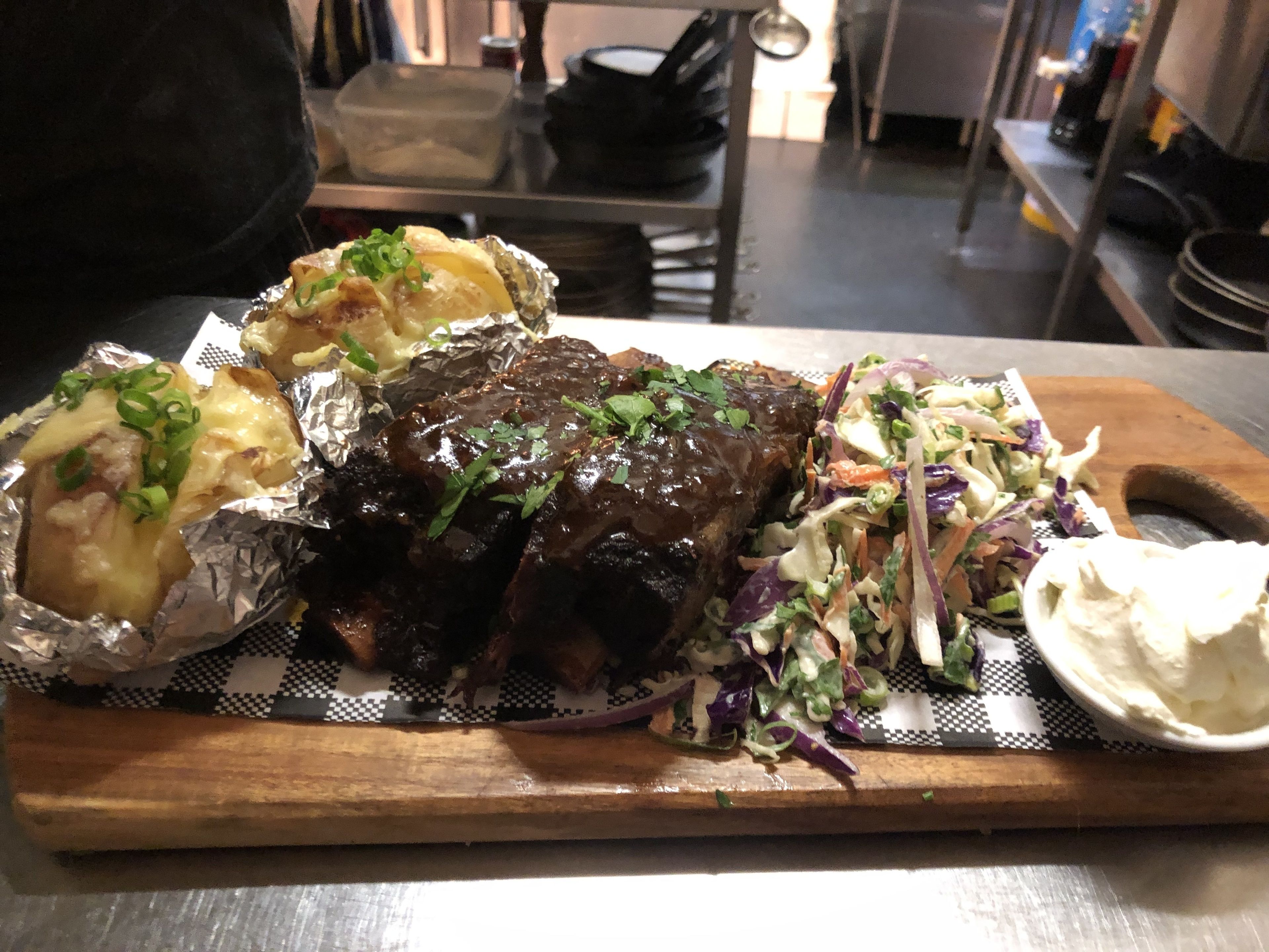 A wooden cutting board with a dish of ribs, foil-wrapped potatoes, and a coleslaw salad.