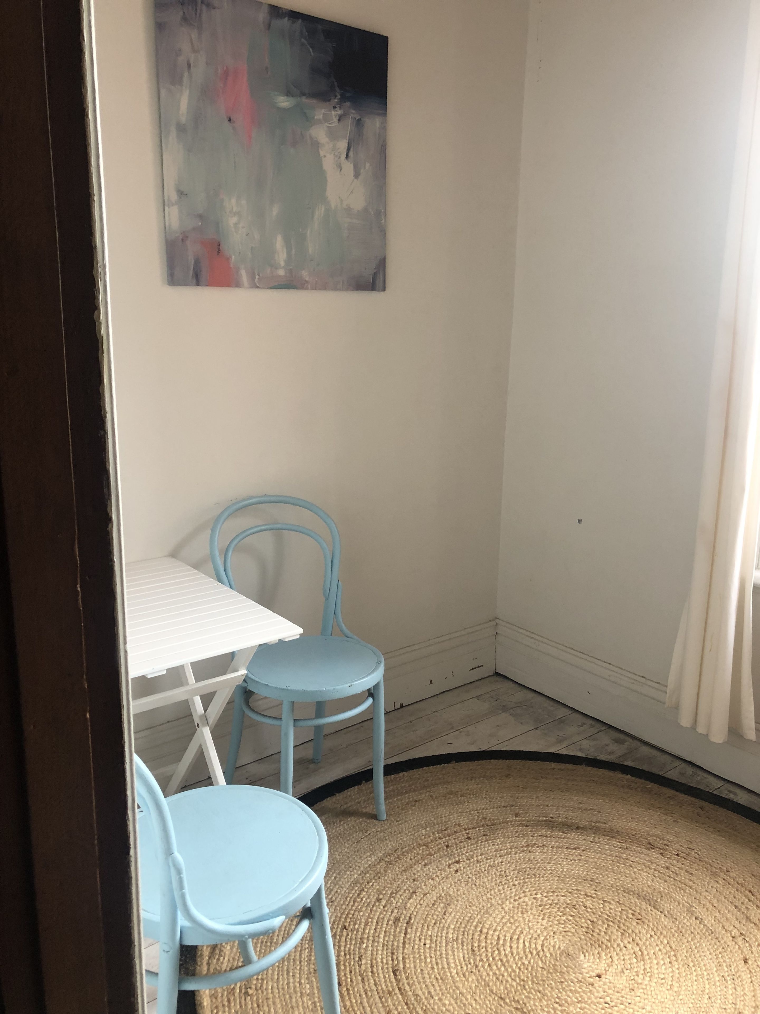 A room with a white table and two blue chairs, a woven rug, and a painting on the wall.