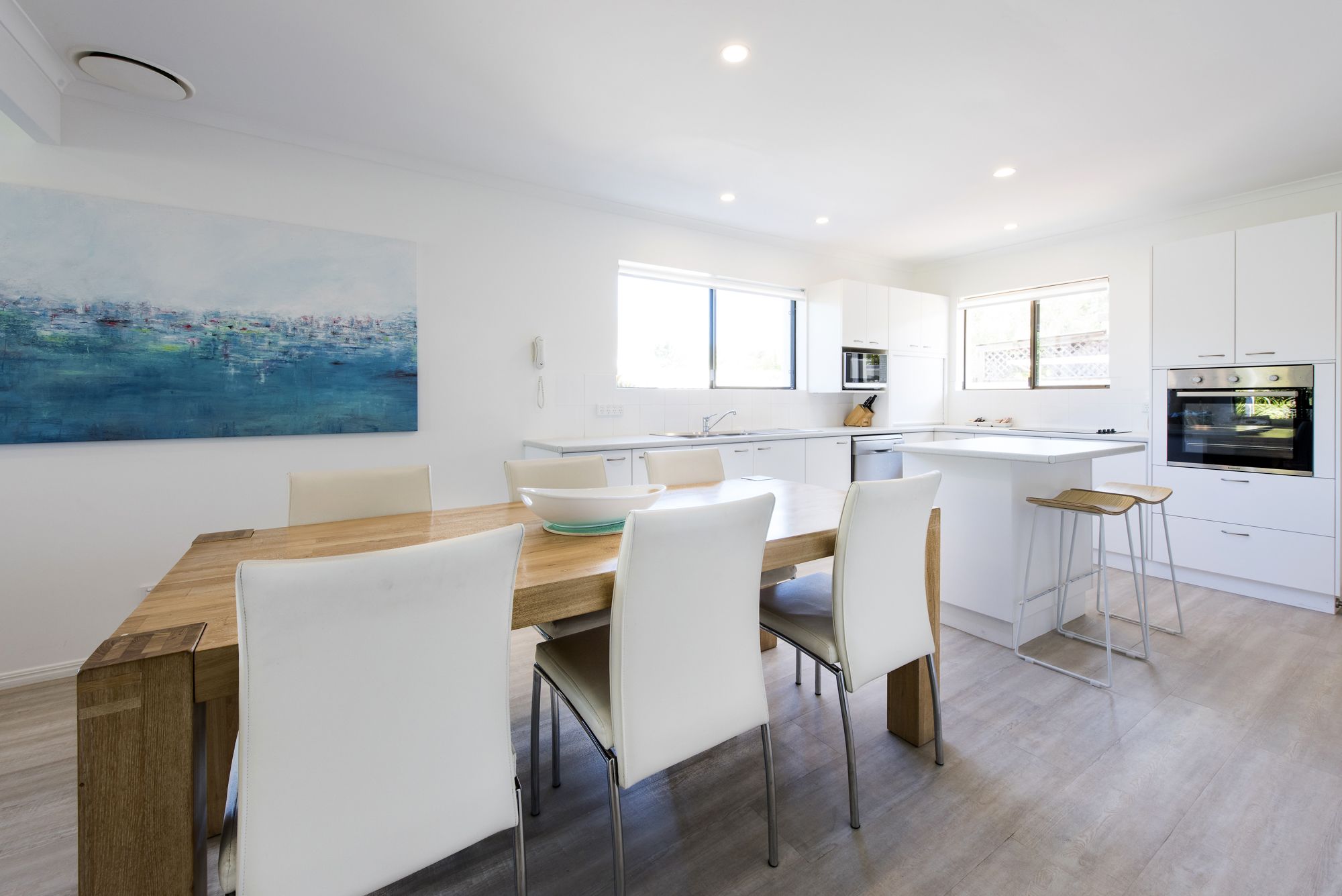 A modern kitchen with white cabinetry, a wooden dining table, and a large painting on the wall.