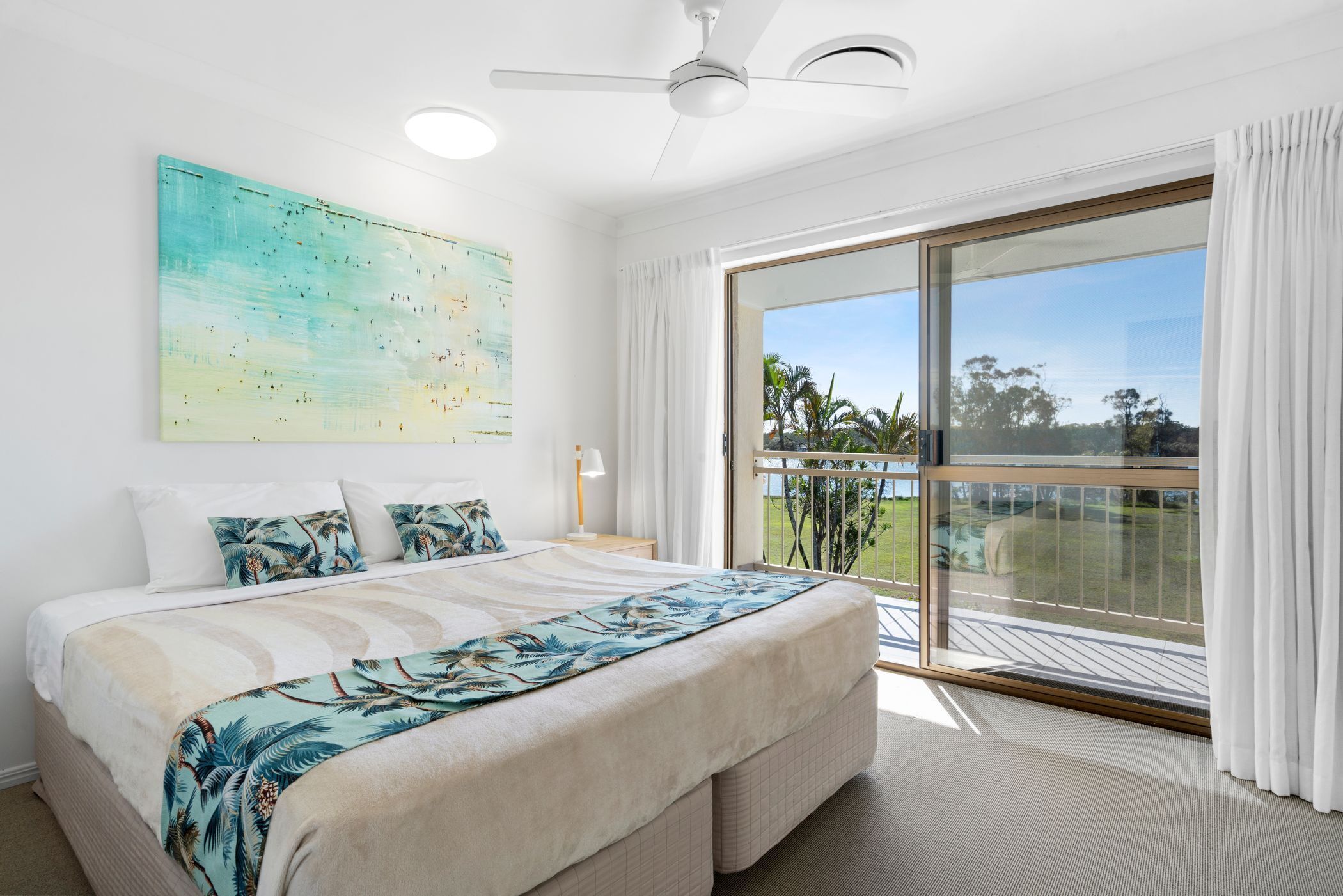A bedroom with a large bed, a ceiling fan, a view of the outdoors, and a painting on the wall.