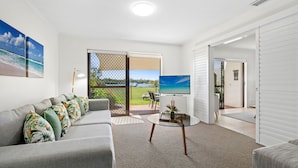 A spacious living room with a grey sofa, a glass coffee table, and a TV. There is a sliding glass door leading to a balcony with a view of a lake and trees. - Cayman Quays (Noosaville)