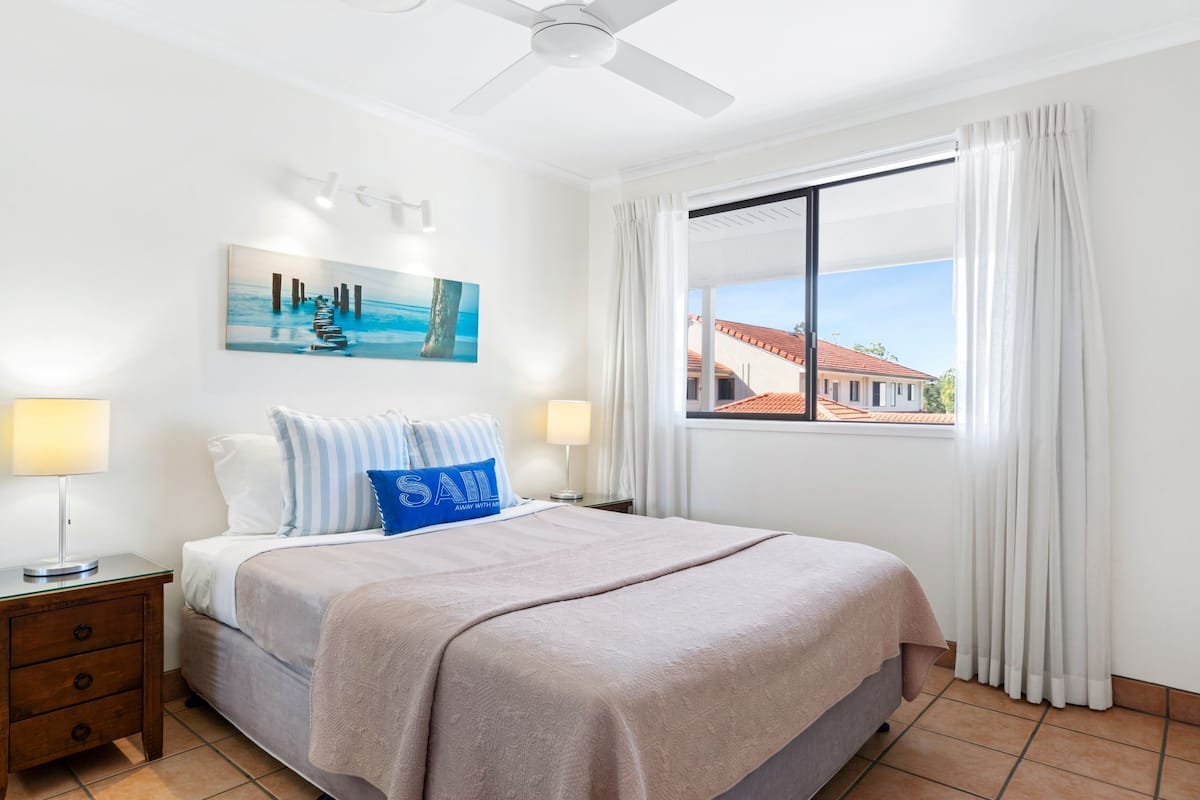 A hotel room with a bed, a nightstand, a lamp, a painting, a window with curtains, and a view of buildings outside.