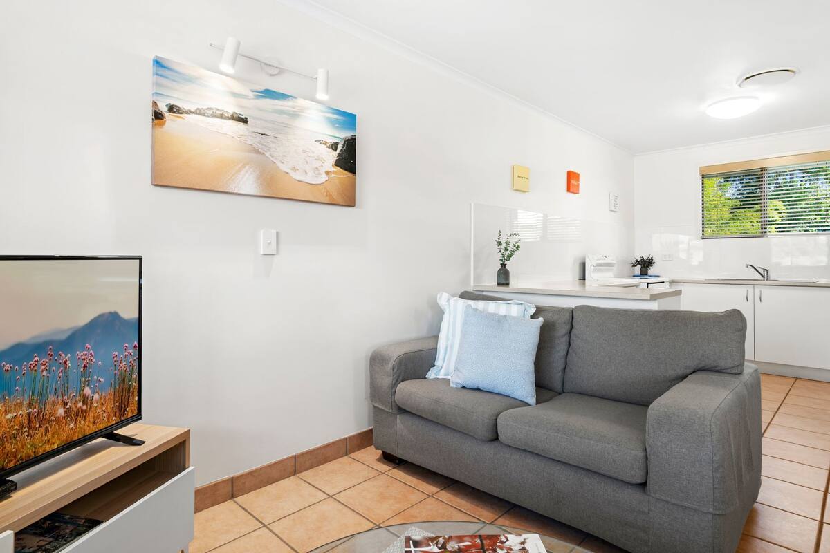 A modern living room with a grey sofa, a flat-screen TV, a coffee table, and a kitchen area in the background.