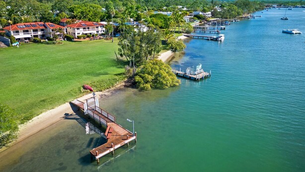 A waterfront property with a dock, a grassy area, and a view of the water and distant hills. - Cayman Quays (Noosaville)