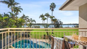 A balcony with a view of a swimming pool, palm trees, and a body of water. - Cayman Quays (Noosaville)