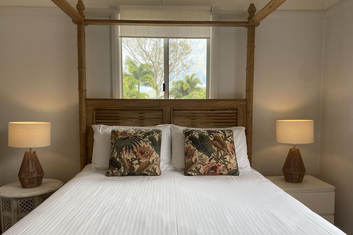 A bedroom with a four-poster bed, two nightstands with lamps, and a window with a view of trees.