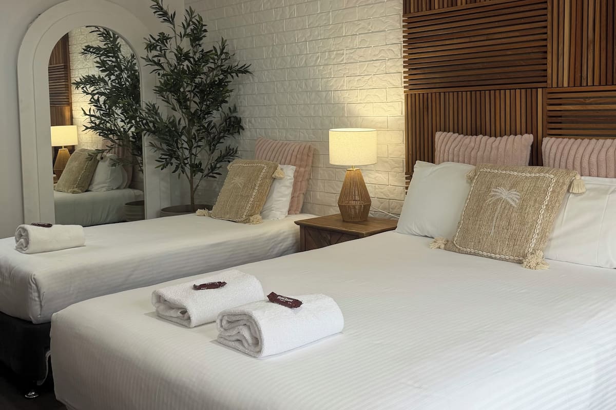 A hotel room with two beds, white linens, a wooden headboard, a large mirror, and a potted plant.