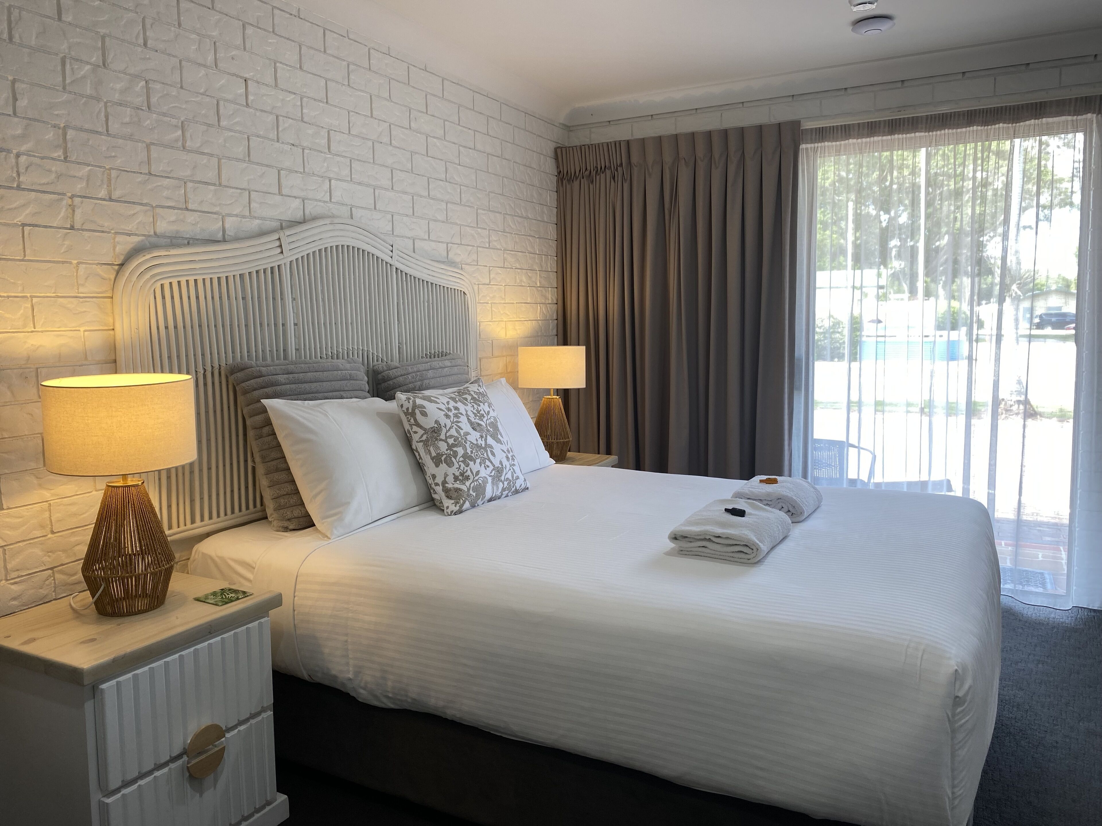 A hotel room with a white brick wall, a bed with white bedding, two bedside lamps, and a view of the outside through large windows.