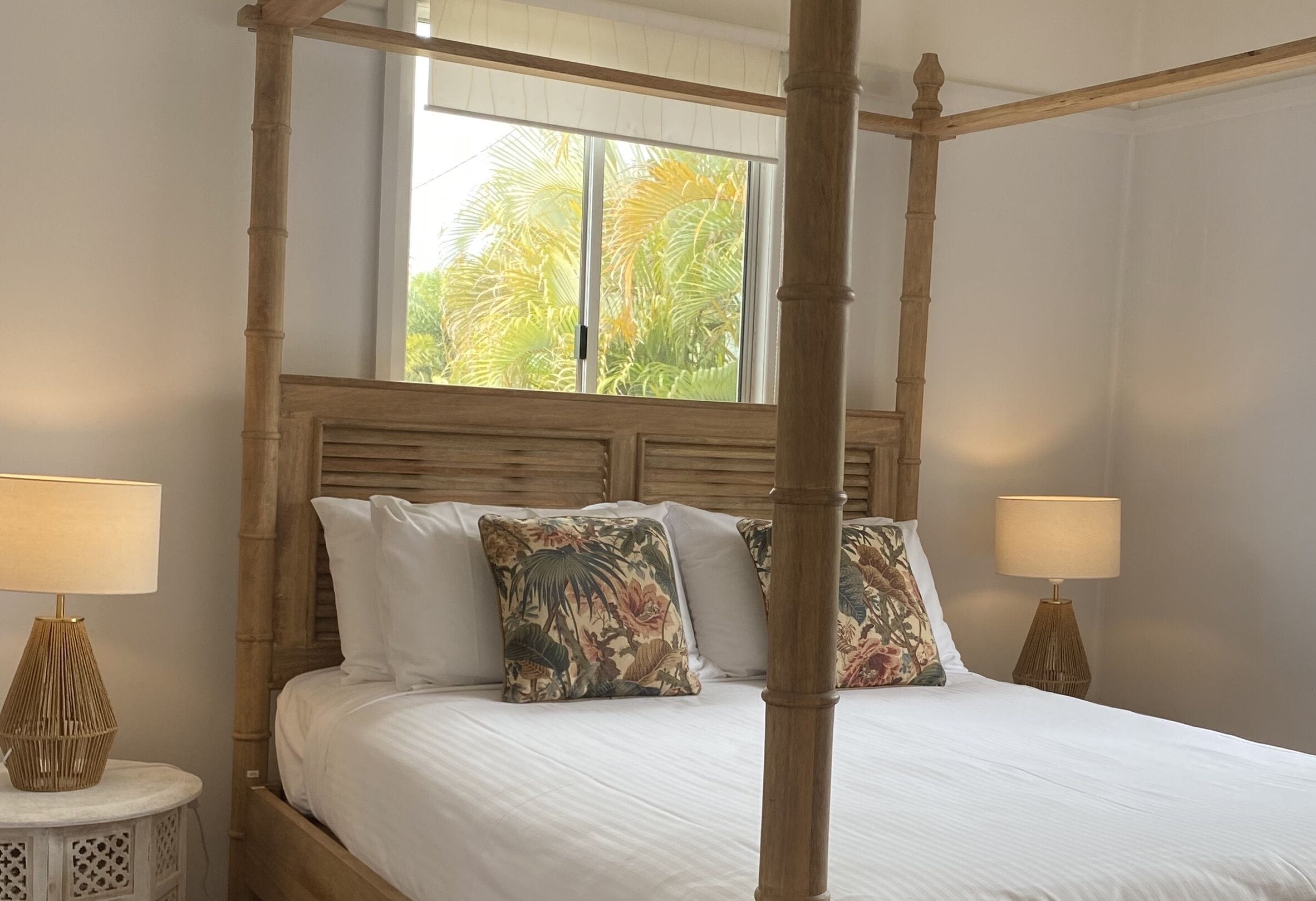 A four-poster bed with white linens and floral pillows, flanked by two bedside tables with lamps, in a room with a large window and a view of palm trees.