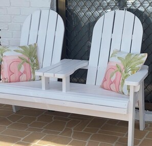 Un banco blanco al aire libre con dos cojines decorados con hojas de palma, situado en un patio con baldosas.