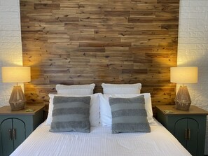 Un dormitorio con cabecera de madera, dos mesitas de noche con lámparas, una cama con ropa blanca y almohadas grises, y armarios verdes.