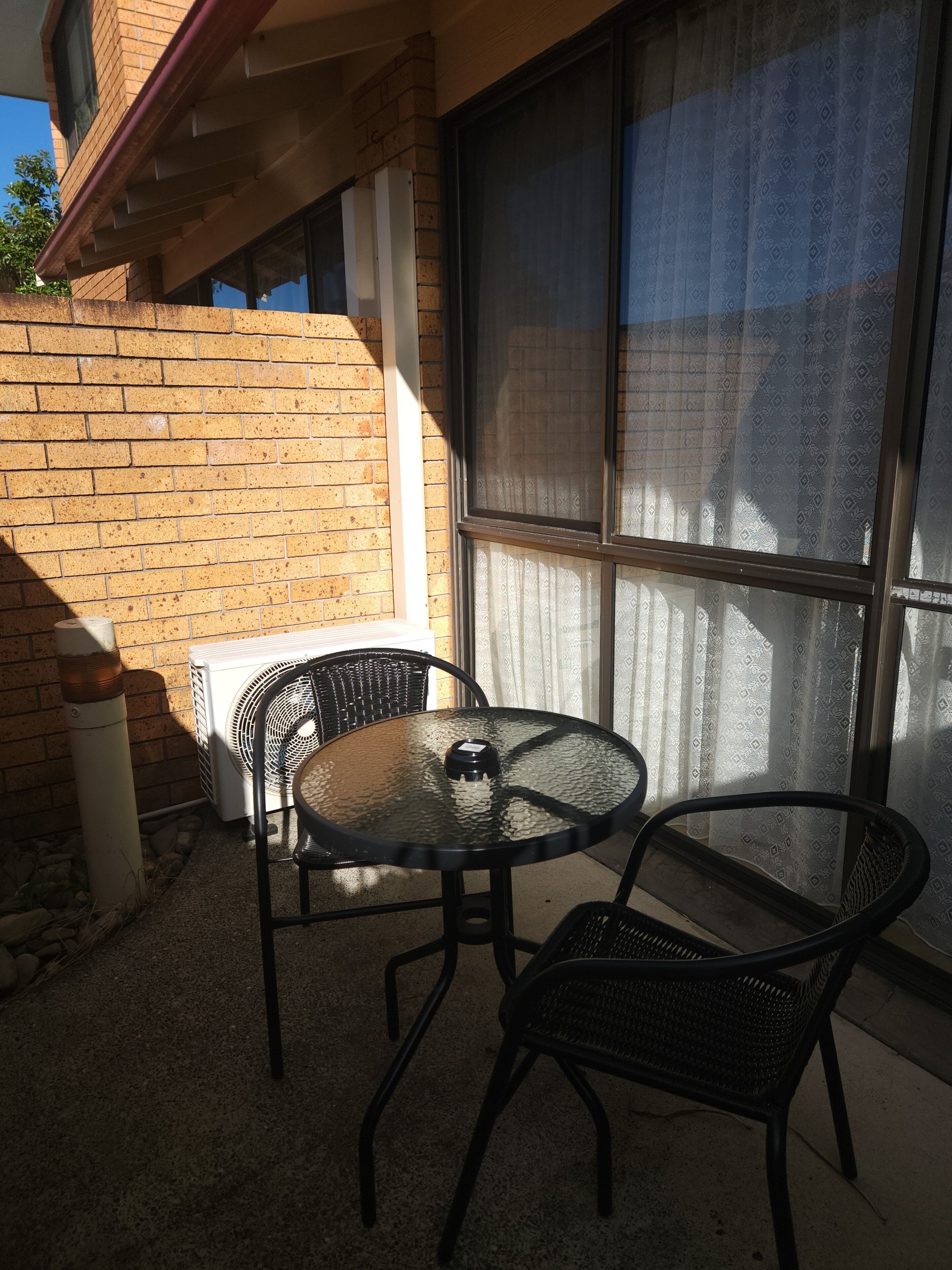 A patio with a round glass table and two chairs, a brick wall, a window with a curtain, and an air conditioning unit.