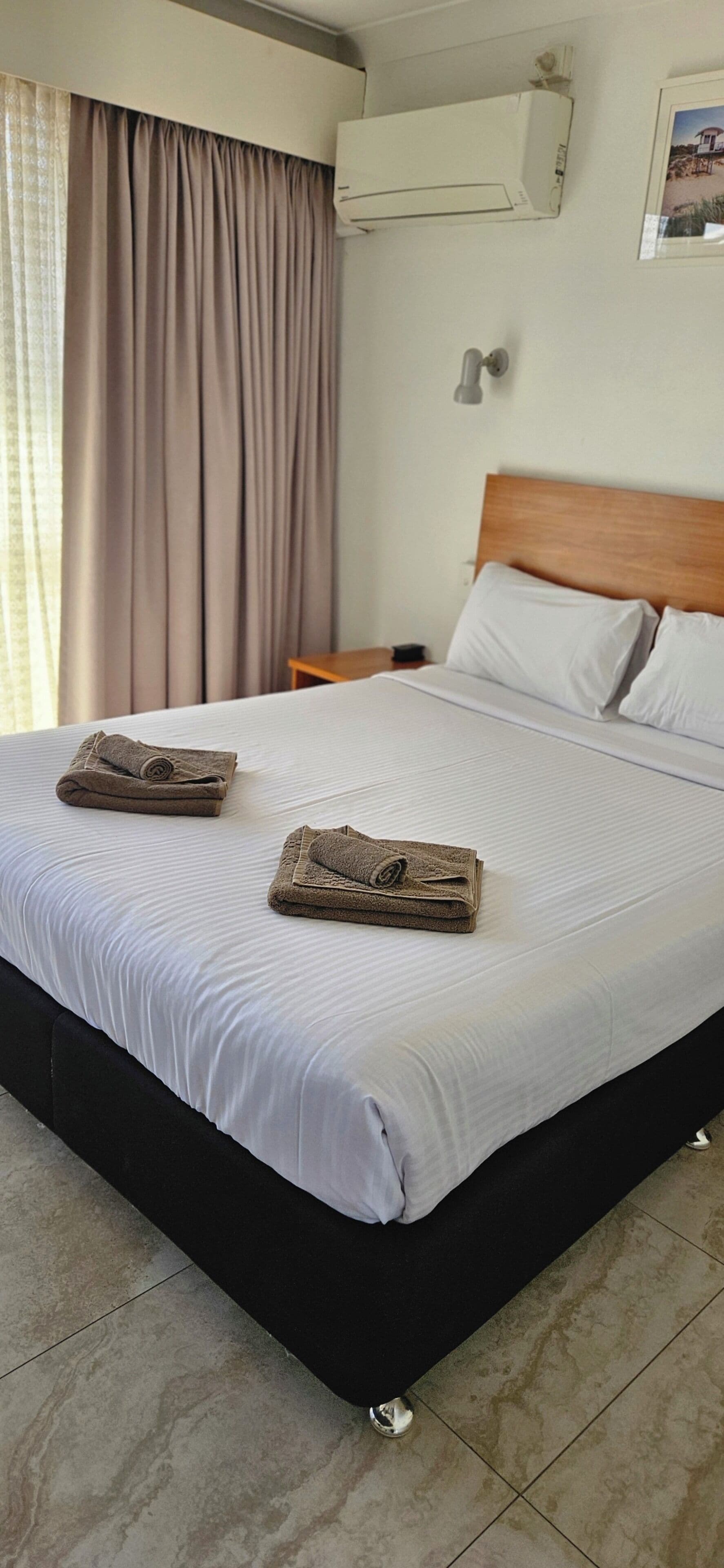 A hotel room with a bed, two towels, a nightstand, a wall-mounted air conditioner, and a window with curtains.
