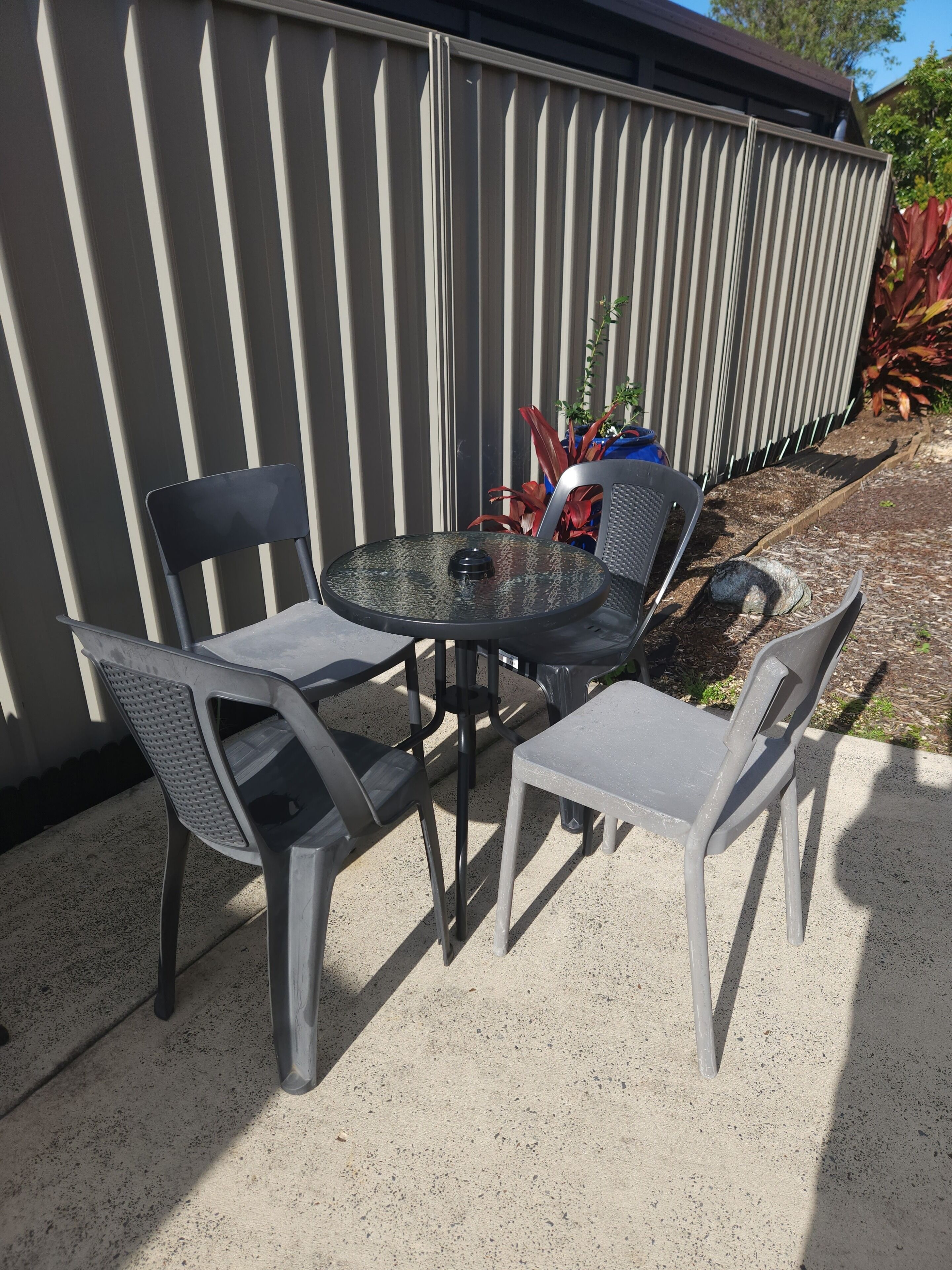 Un patio al aire libre con una mesa redonda y cuatro sillas, una planta en maceta y una cerca metálica al fondo.
