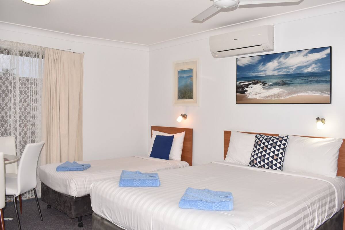 A hotel room with two beds, a desk, and a chair. There is an air conditioning unit on the wall and a painting of a beach scene.