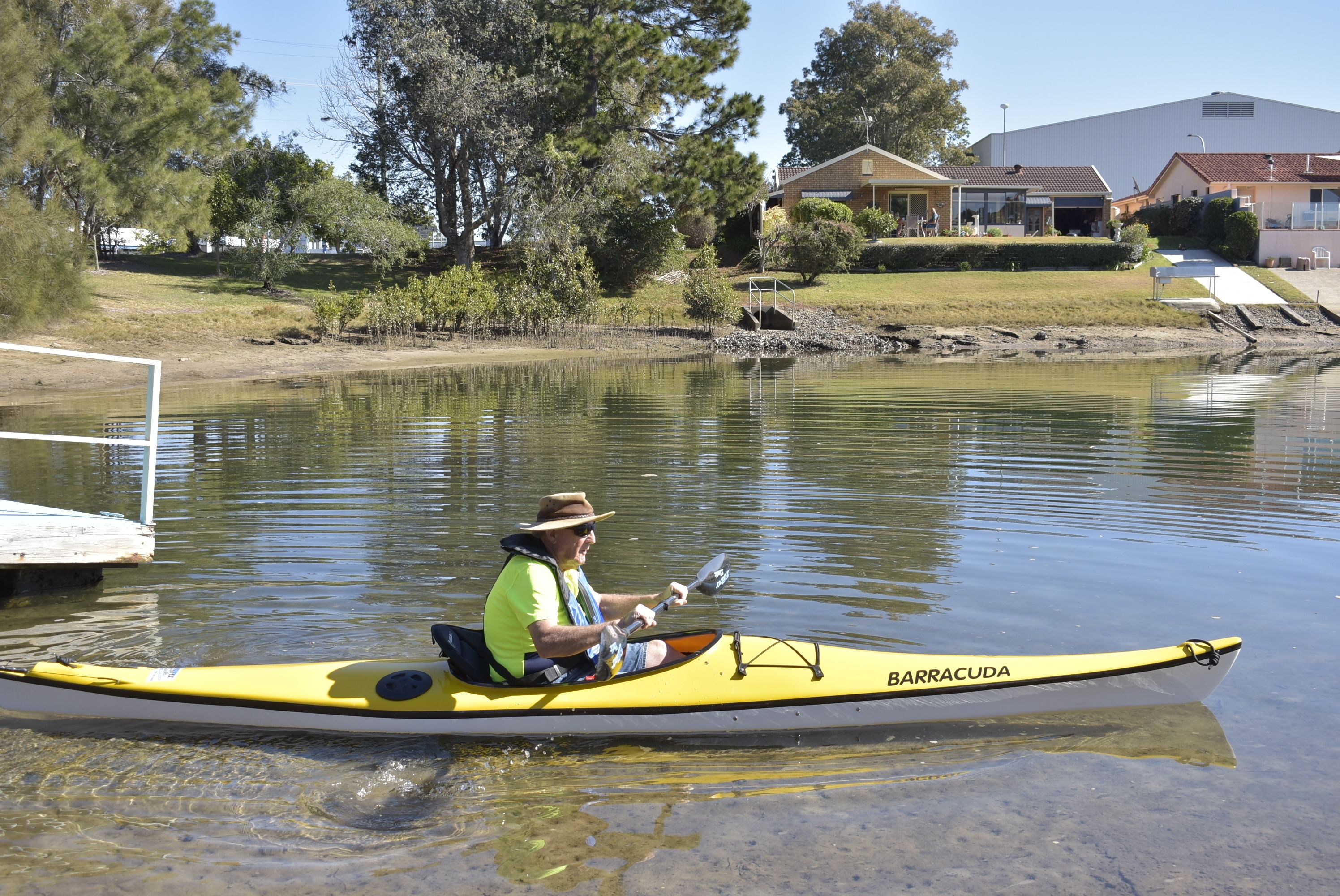 A man in a yellow kayak labeled "BARRACUDA" on a calm lake with a dock in the background and houses nearby.