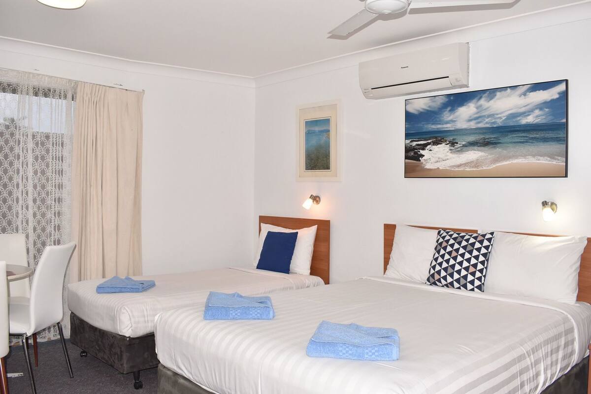 A hotel room with two beds, a desk, a chair, an air conditioner, and a painting of a beach.