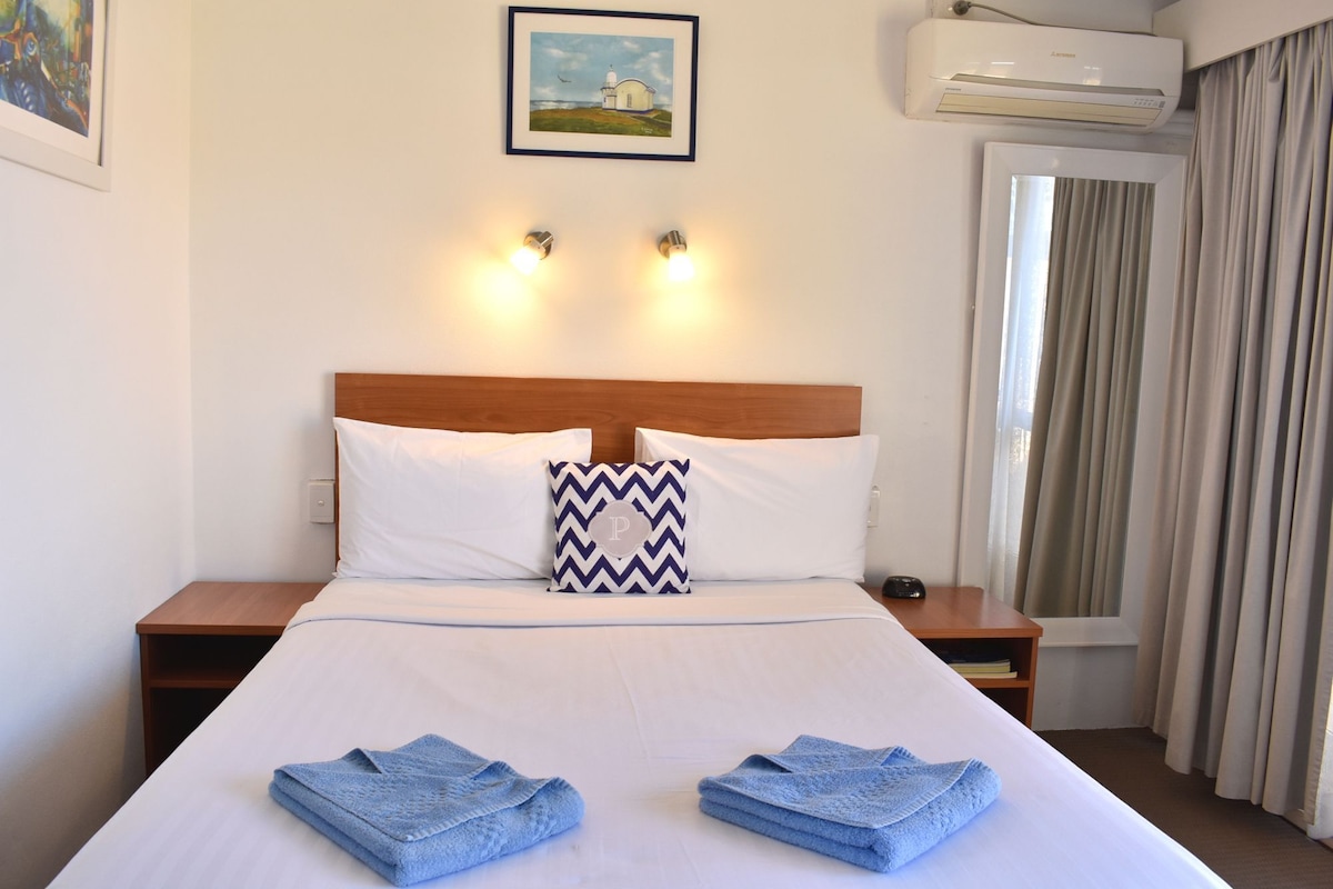 A hotel room with a bed, two bedside lamps, a picture on the wall, an air conditioner, and a window with curtains.