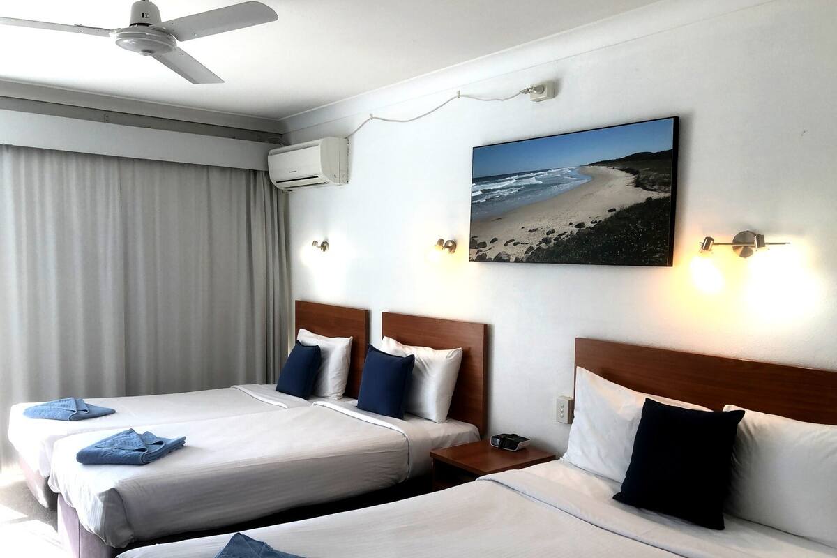 A hotel room with two beds, a ceiling fan, an air conditioning unit, a painting of a beach, and a bedside table with a lamp.