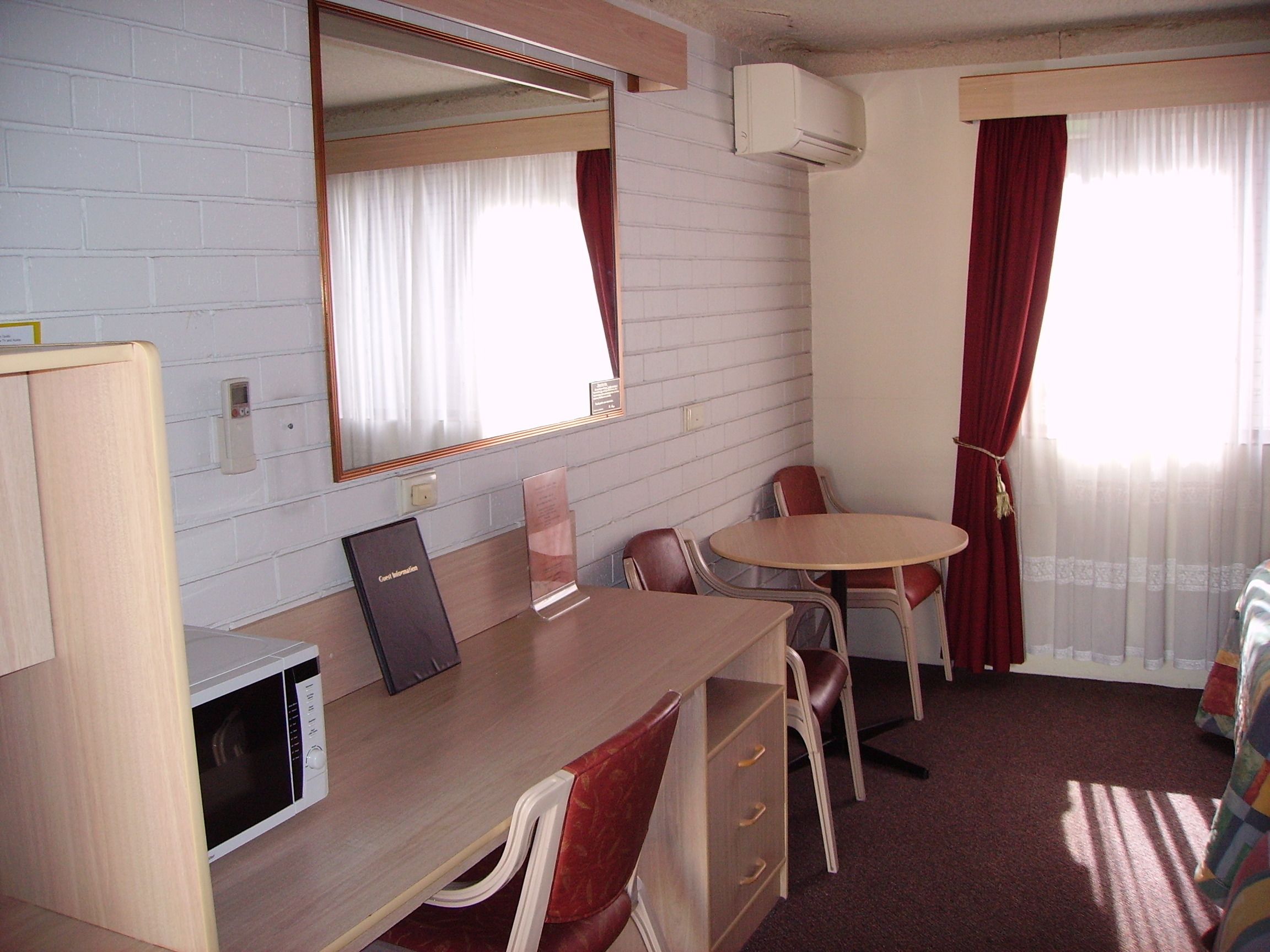 A compact hotel room with a wooden desk, a chair, a small table, a microwave, a mirror, and a window with curtains.