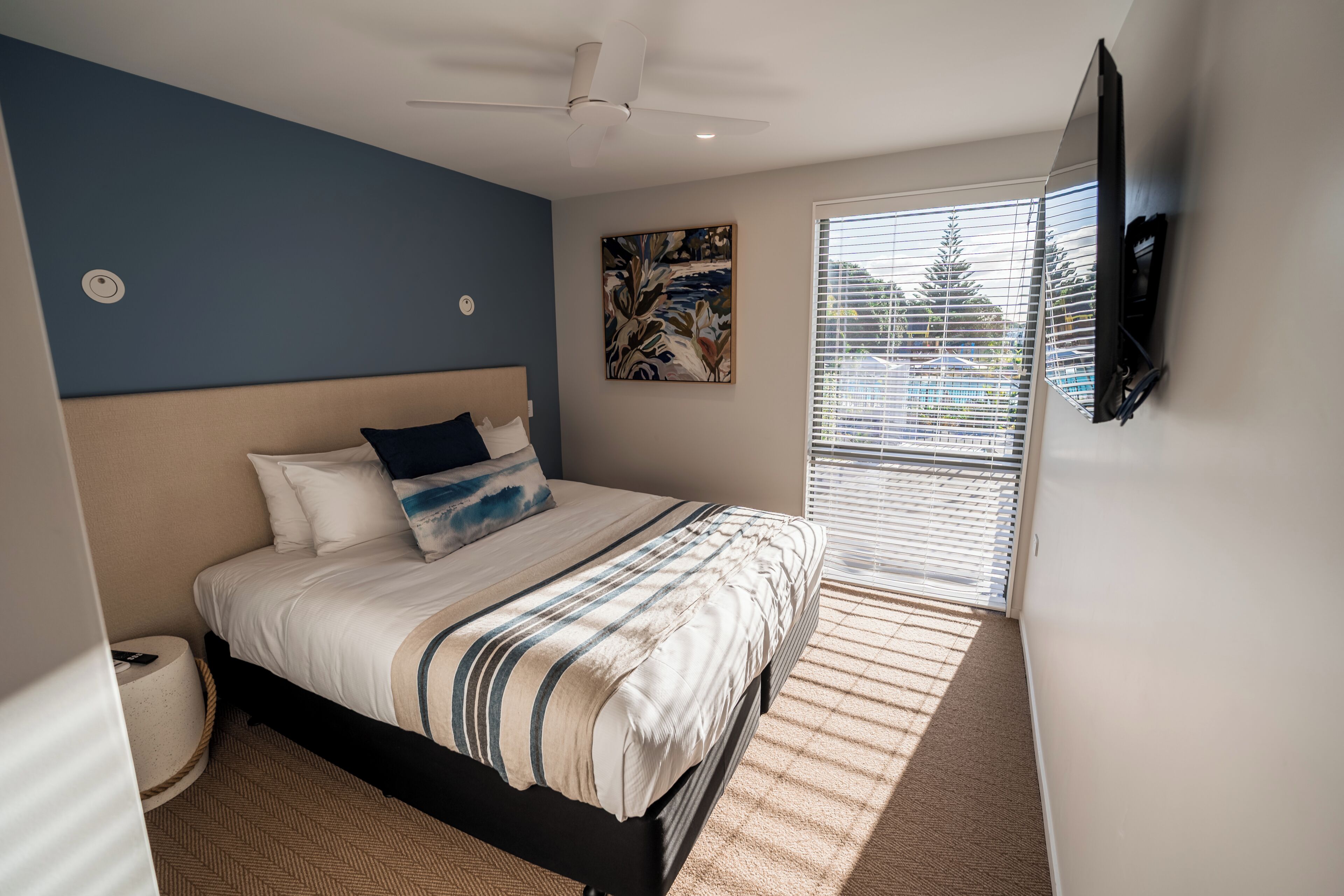 A bedroom with a bed, a ceiling fan, a painting, a window with blinds, and a TV mounted on the wall.