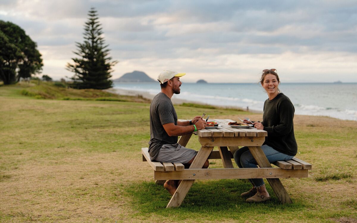 Dos personas sentadas en una mesa de picnic de madera en un campo cubierto de hierba, con un cuerpo de agua y árboles al fondo.