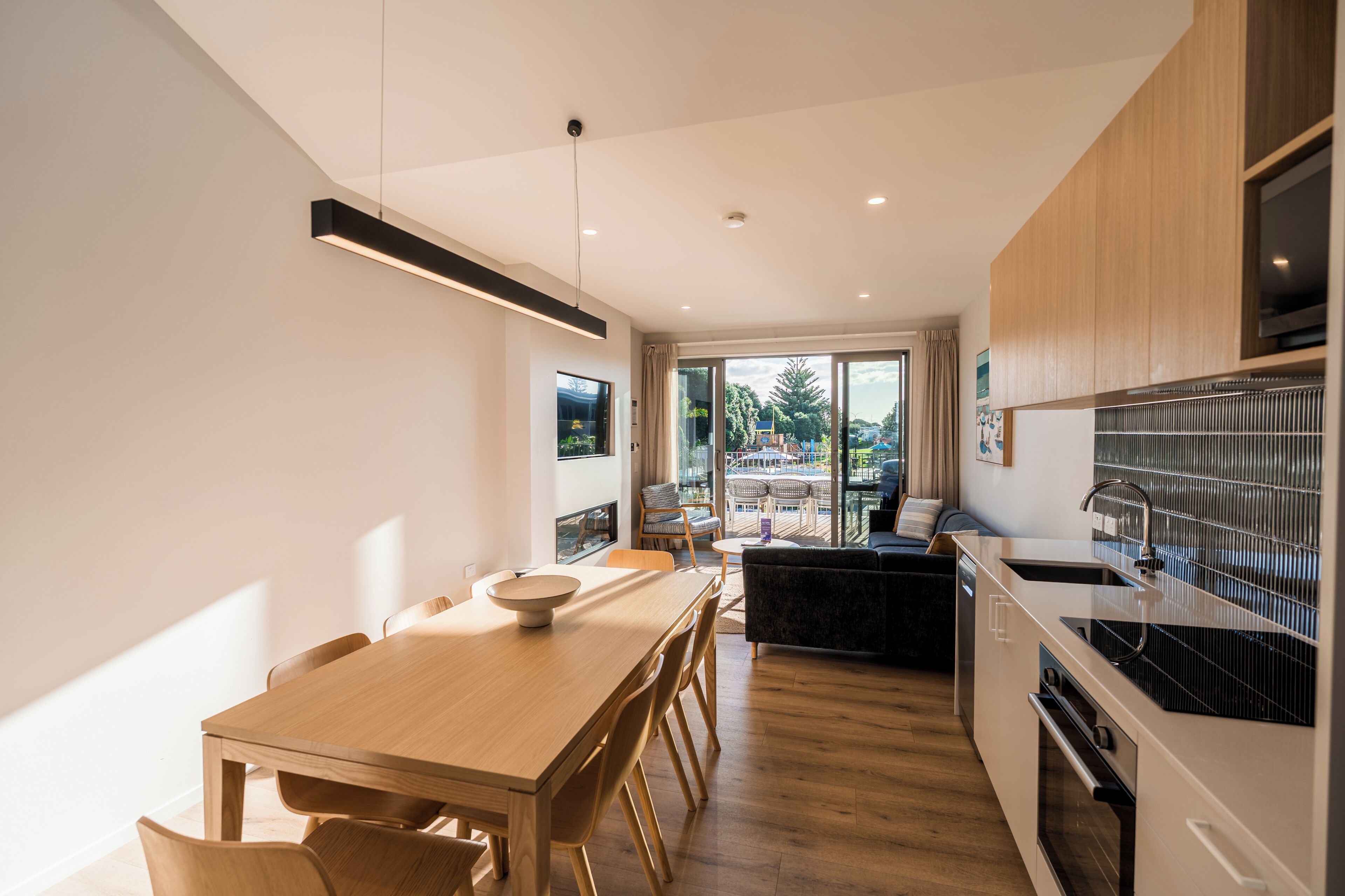 Een moderne keuken met houten kastjes, een eethoek met een tafel en stoelen, en uitzicht door de glazen deur naar buiten.