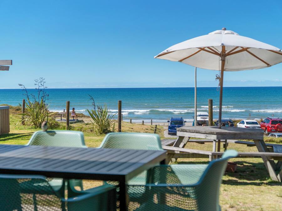 Un patio frente a la playa con mesas y sillas, una pérgola de madera y vistas al océano.