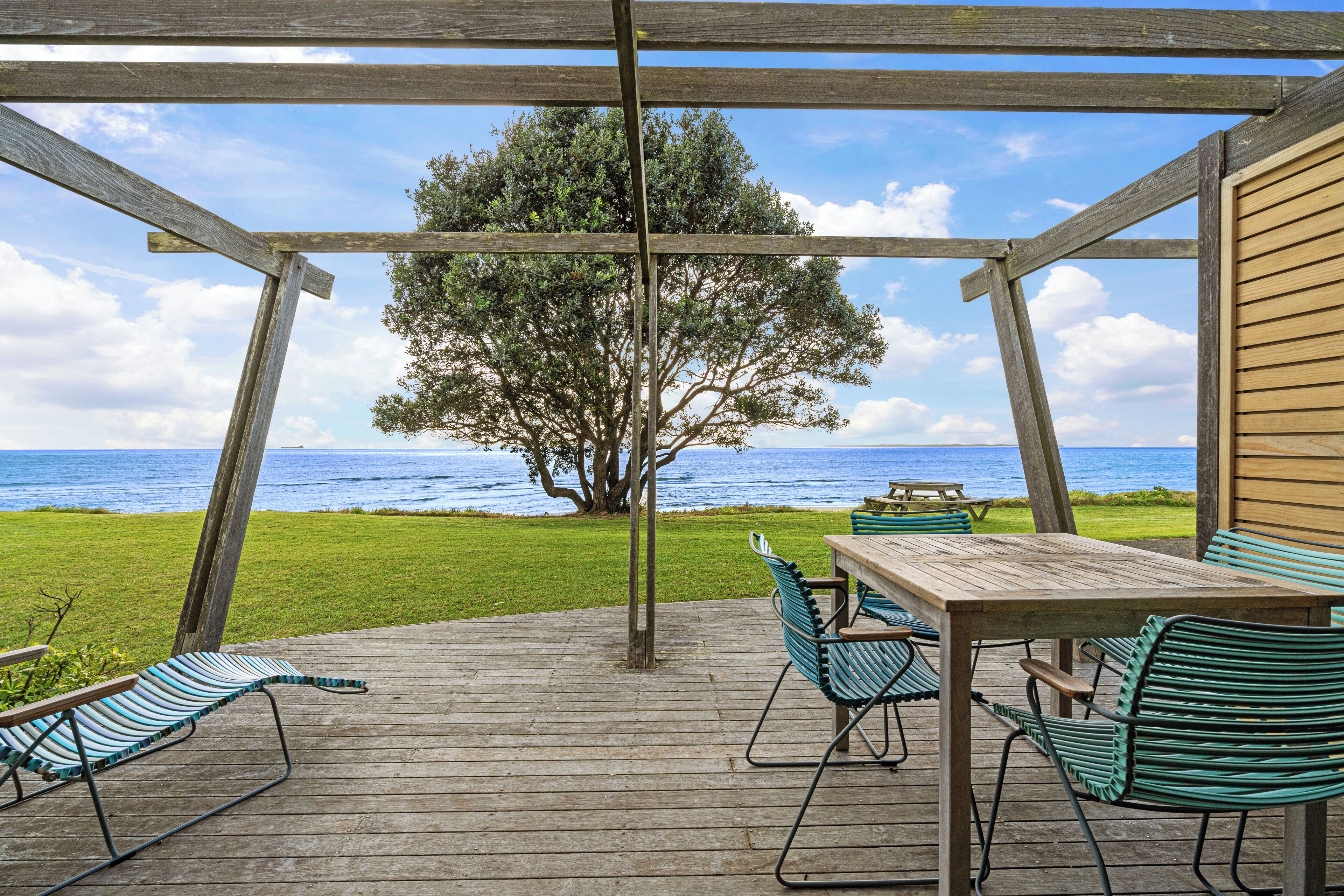Una terraza de madera con una mesa y sillas, con vista a un área verde y al océano.