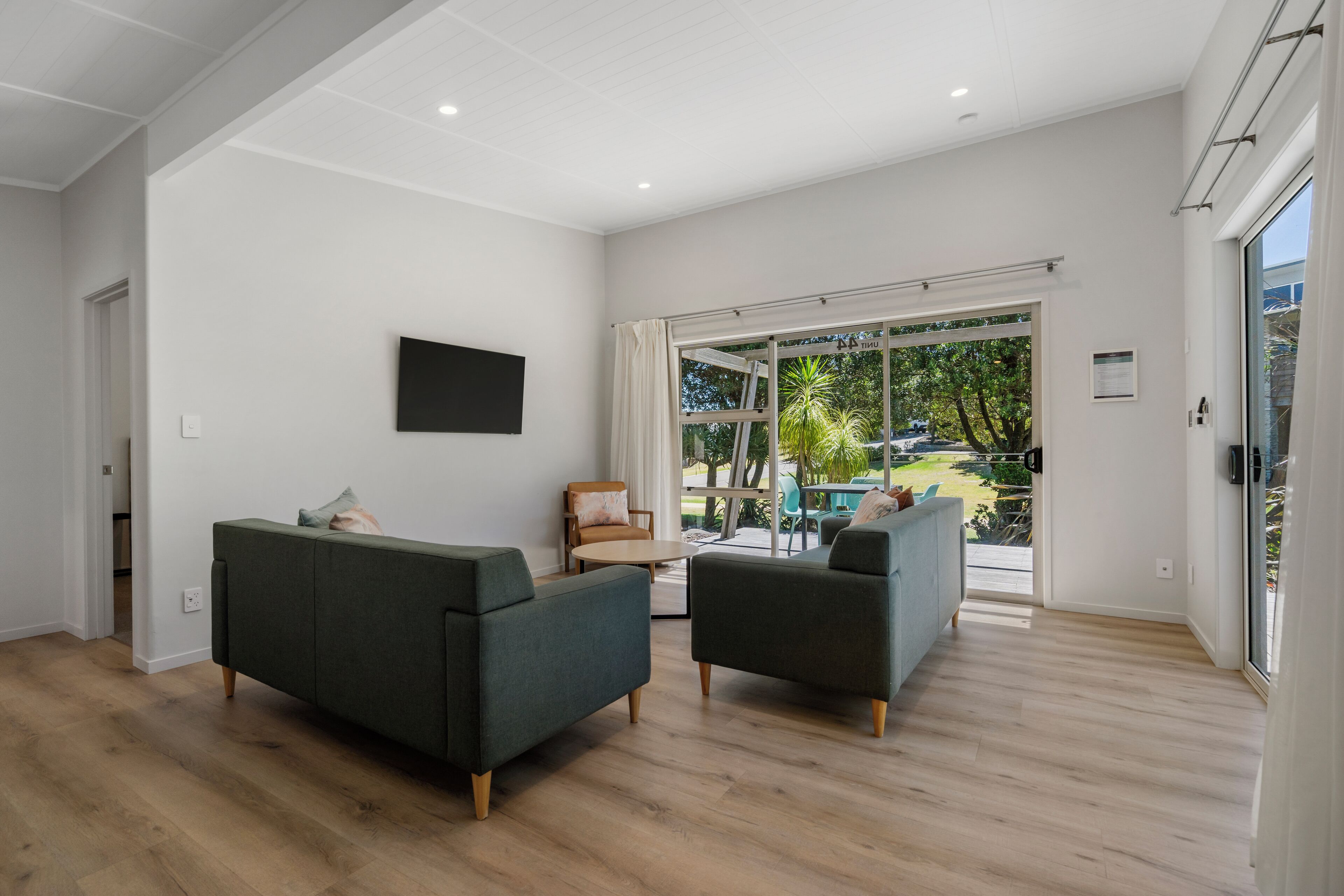 A spacious living room with a large sliding glass door leading to a balcony, a flat-screen TV mounted on the wall, and a wooden floor.