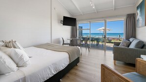 A bedroom with a bed, a sofa, and a TV. There is a view of the beach through the window.