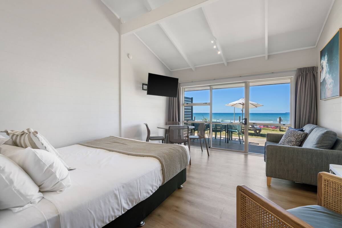Ein Schlafzimmer mit einem Bett, einem Sofa und einem Fernseher. Durch das Fenster ist der Strand zu sehen.