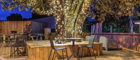 An outdoor patio area with wooden tables and chairs, a wooden deck, and a tree decorated with lights.