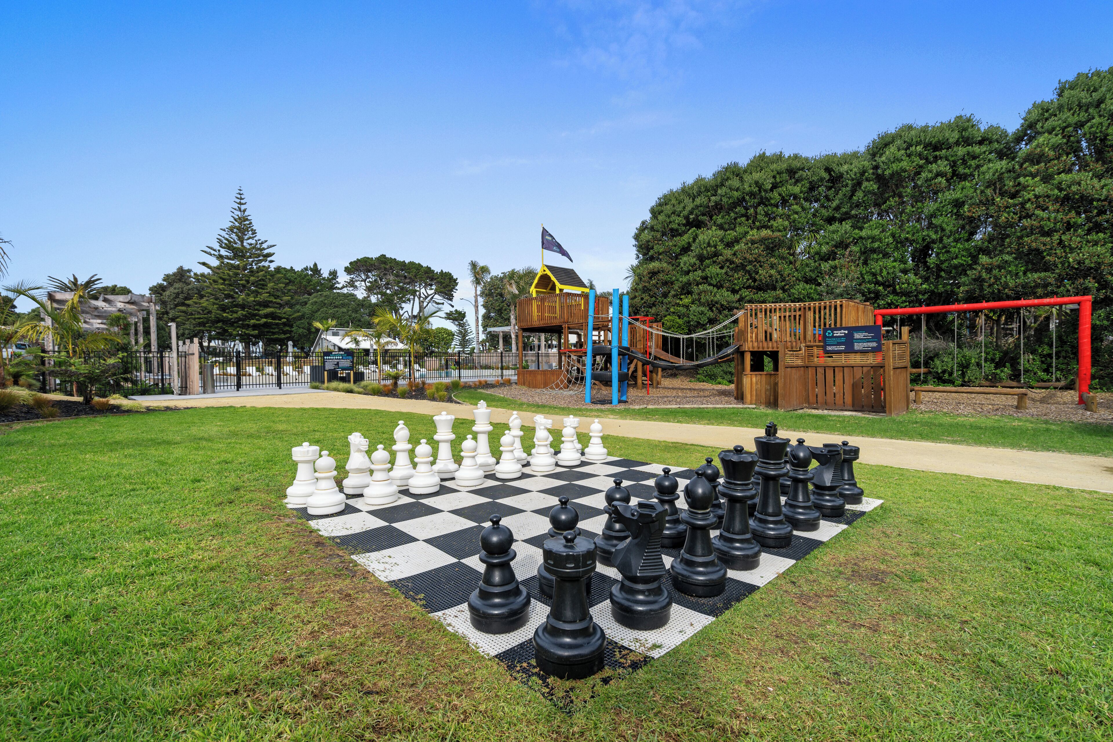 Un tablero de ajedrez grande al aire libre sobre un césped, con un parque infantil y estructuras de madera al fondo.