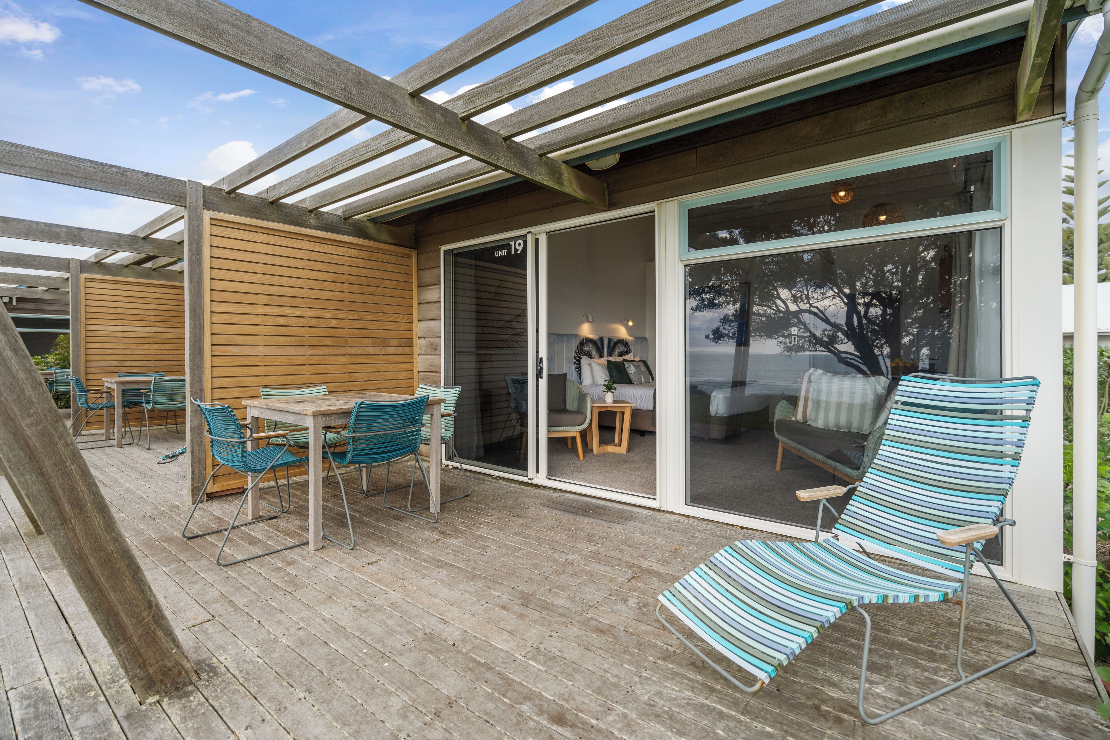 Una terraza de madera con mobiliario exterior, una puerta corrediza de vidrio que da a una habitación y una vista despejada del exterior.