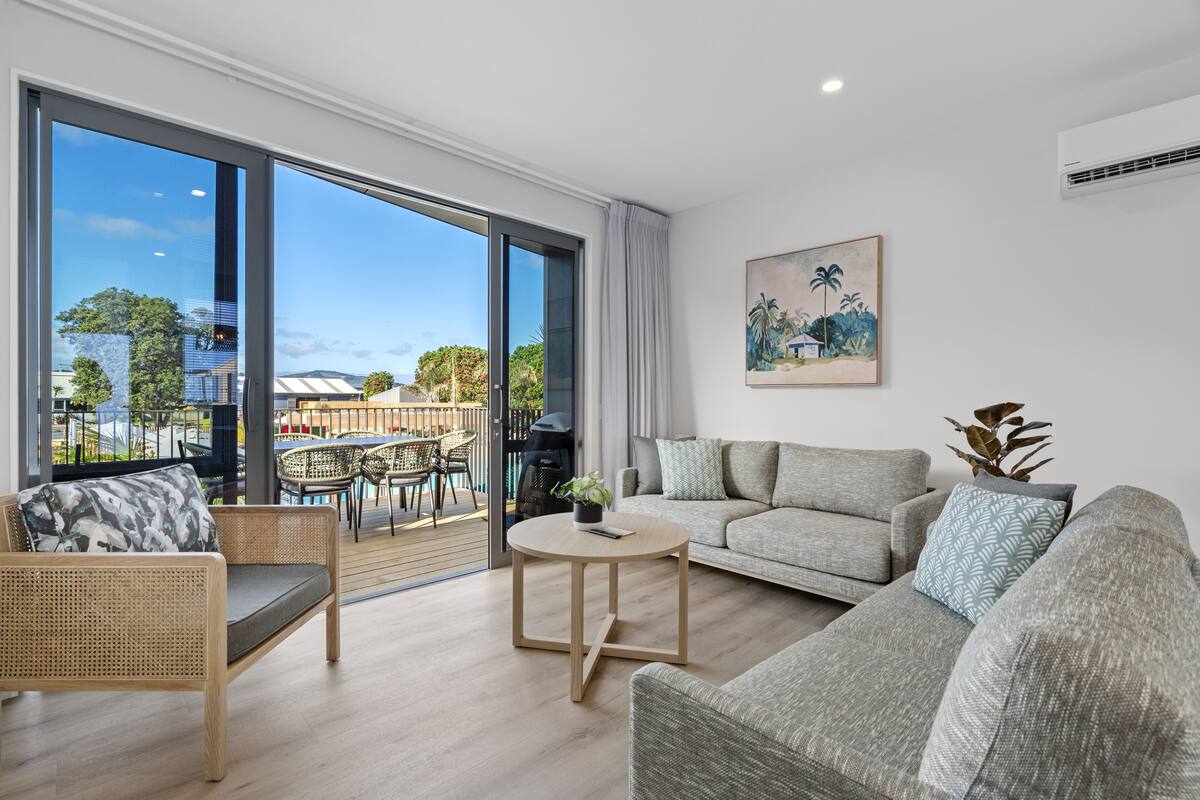 Ein modernes Wohnzimmer mit einer Couch, einem runden Couchtisch und einem Korbstuhl. Ein großes Fenster bietet einen Blick ins Freie.