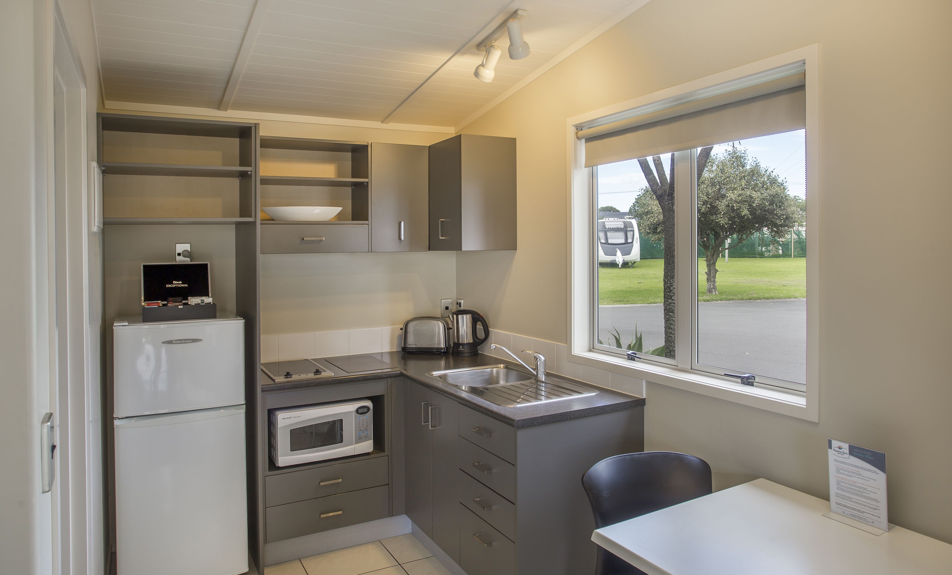 A compact kitchen with a refrigerator, microwave, toaster, and sink. There is a window with a view of a parking area and trees.