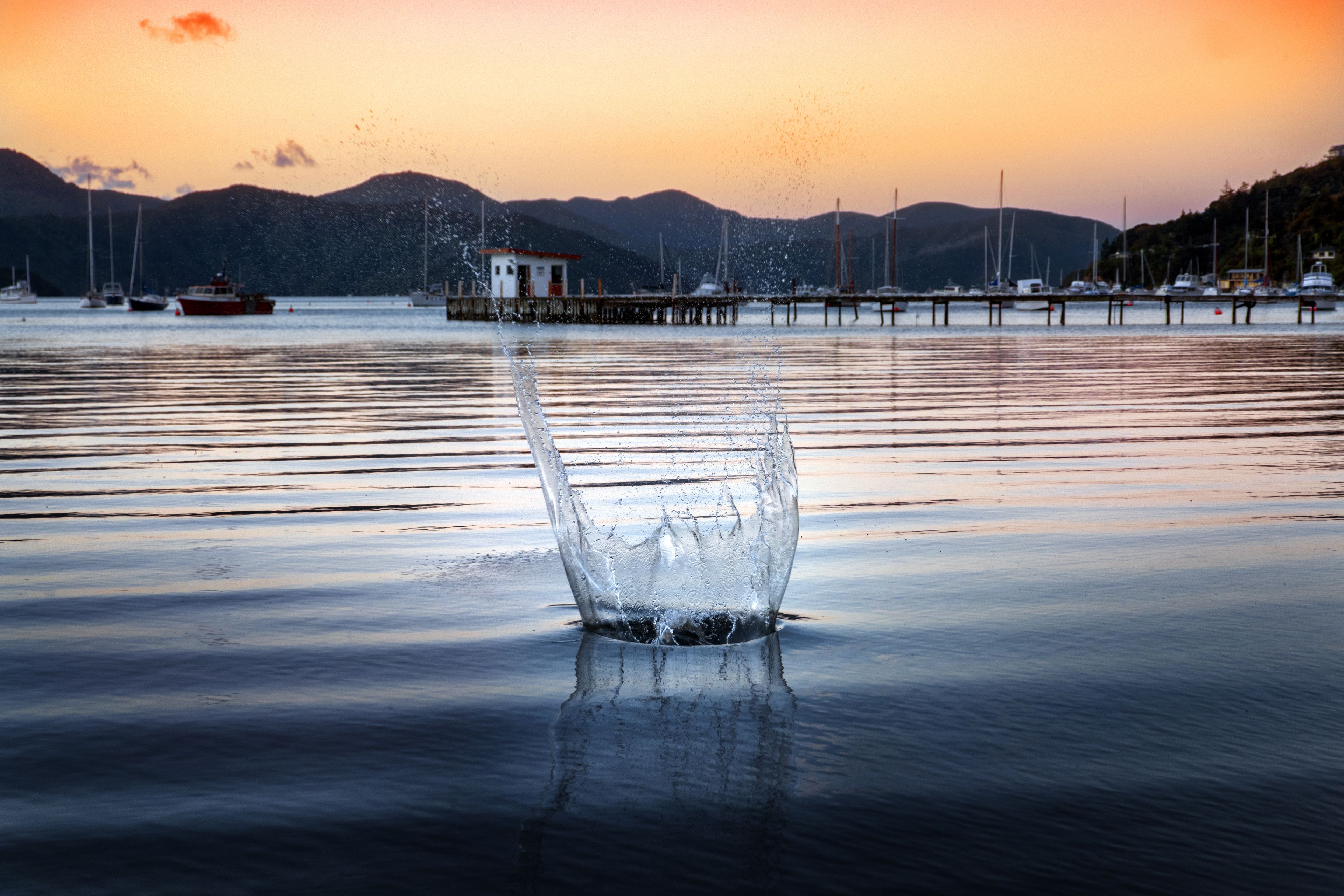 A calm body of water with a splash in the foreground, boats, and a pier in the background.
