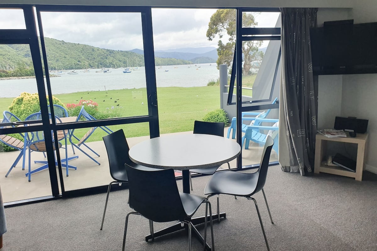 Une pièce dotée d’une grande fenêtre donnant sur un lac et des collines, comprenant une table ronde avec des chaises, un téléviseur posé sur un meuble et une petite table d’appoint.