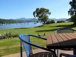 A lakeside view with a wooden table and blue metal chairs, ducks on the grass, and boats on the water. - Bay Vista Waterfront Motel Picton (Picton)