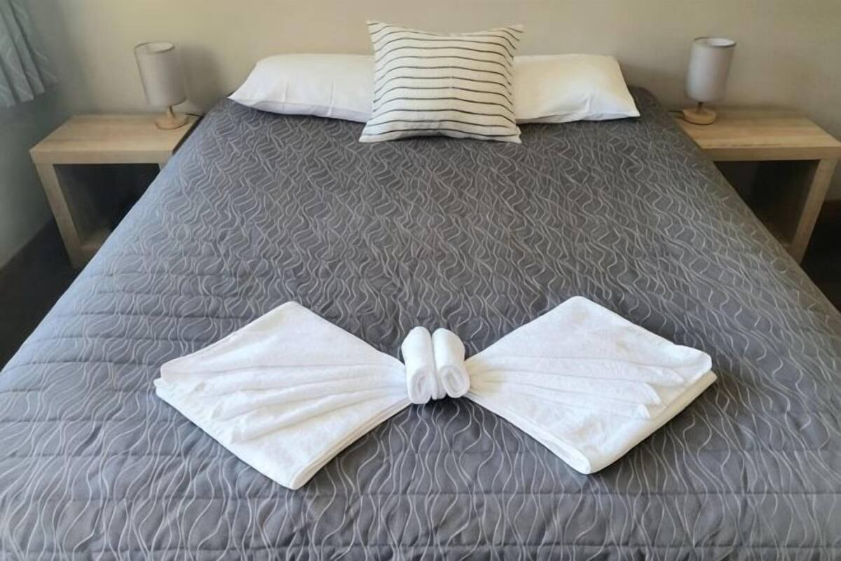 Un lit bien fait, avec une couverture grise, deux oreillers et une table de chevet sur laquelle se trouve une lampe.