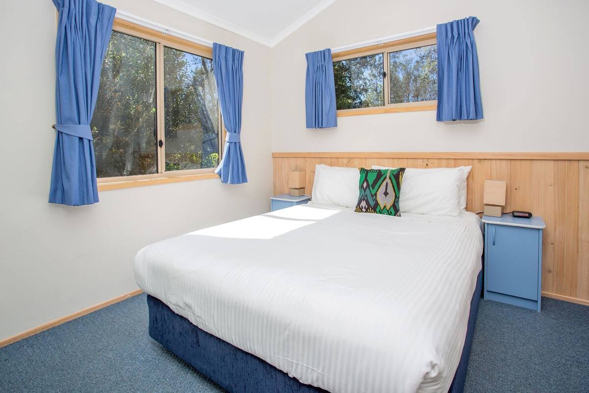 A bedroom with a bed, two nightstands, a wooden headboard, and two windows with curtains.