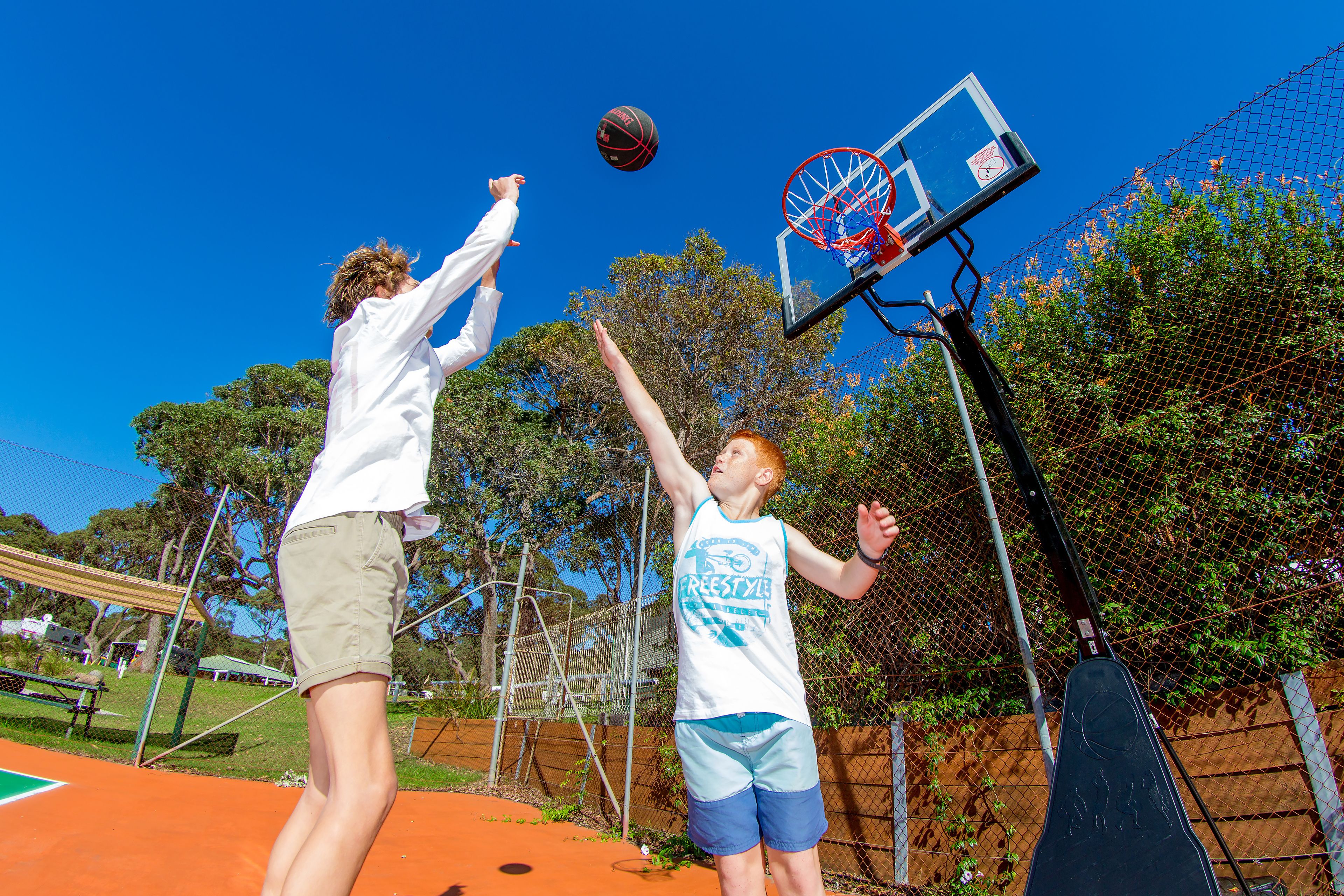 Dos personas jugando baloncesto en una cancha al aire libre con una cerca de malla y árboles al fondo.