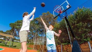 Dos personas jugando baloncesto en una cancha al aire libre con una cerca de malla y árboles al fondo.