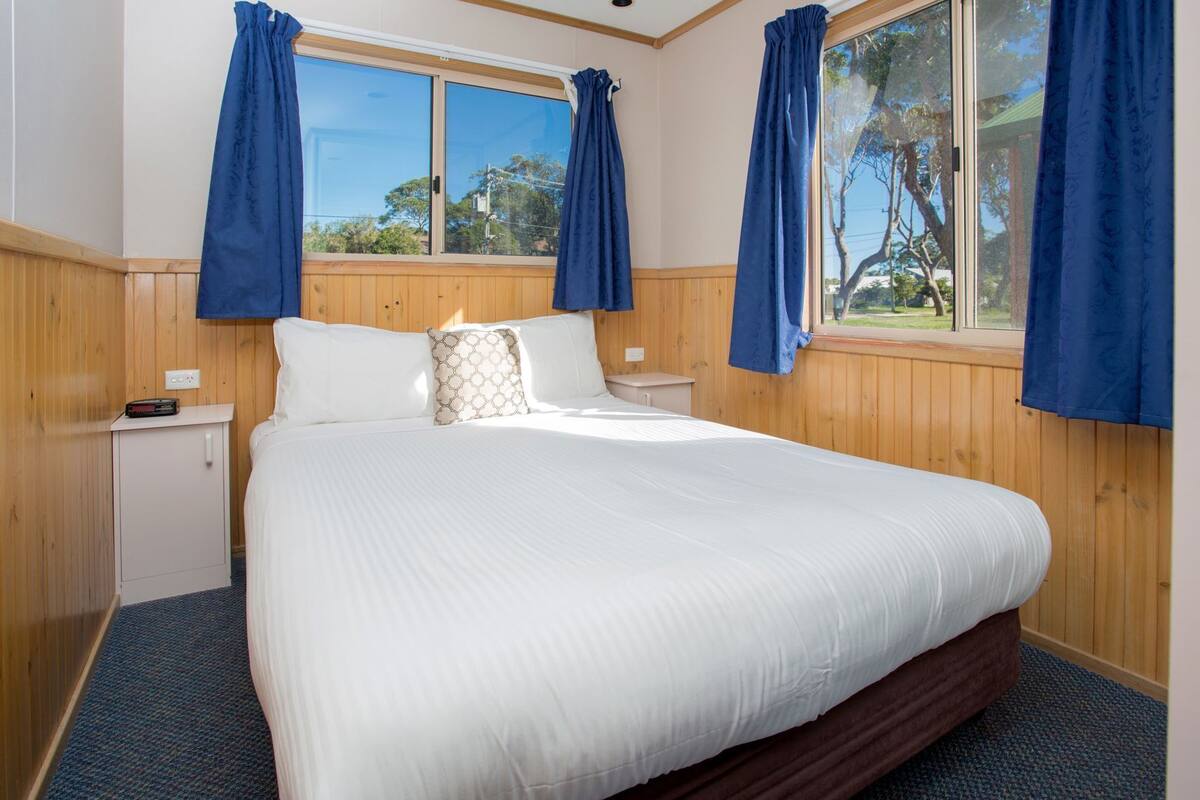A bedroom with a large bed, wooden walls, a nightstand, and a window with curtains.