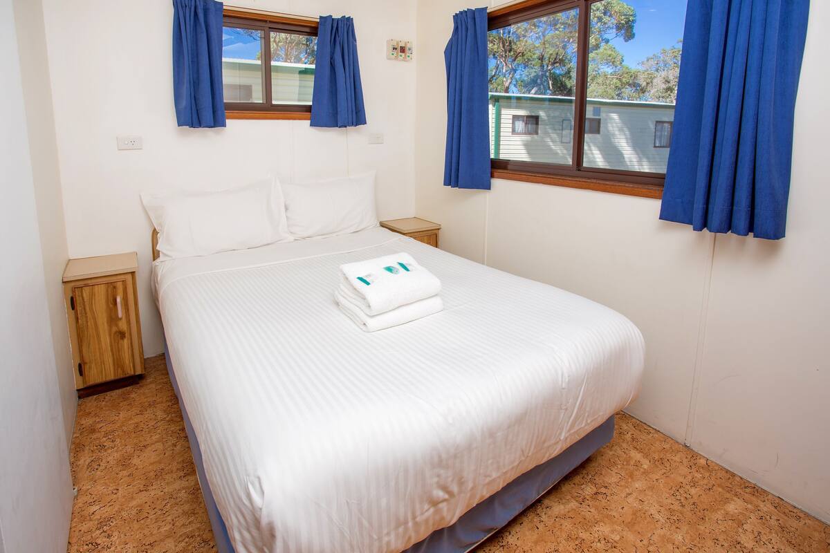 A neatly made bed with white linens and a folded towel, wooden nightstands, and blue curtains on a window.