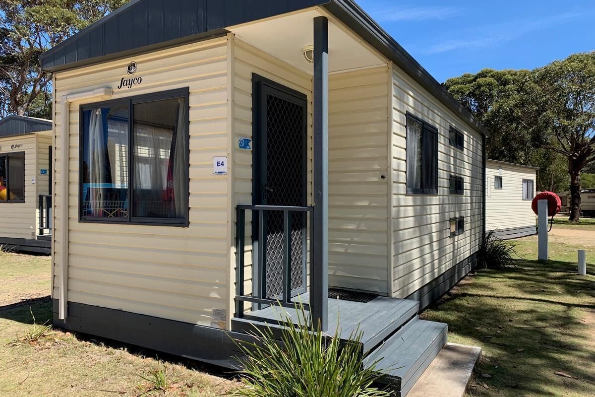 A mobile home with a front porch, a window with curtains, and a small outdoor area with grass and plants.