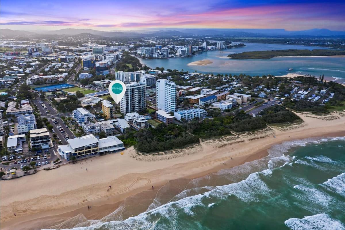 An aerial view of a coastal city with a prominent hotel marked by a location pin, surrounded by residential buildings, a beach, and a bay.